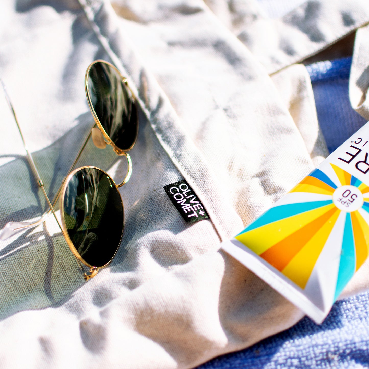 An Olive+Comet Sonoma bag lies on the beach. Sitting on a blue & white striped towel, the bag is surrounded by aviator-style sunglasses and reef-friendly sunscreen.