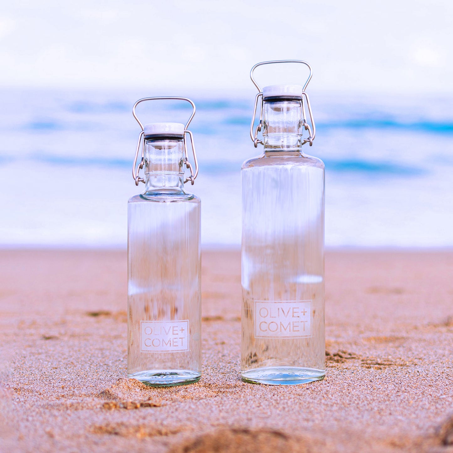 Two Hadley bottles stand side-by-side at the ocean's edge. On the left is the .6 liter variant—and on the right, the 1.0 liter version for comparison. The bottles are identical looking, except for the capacity.