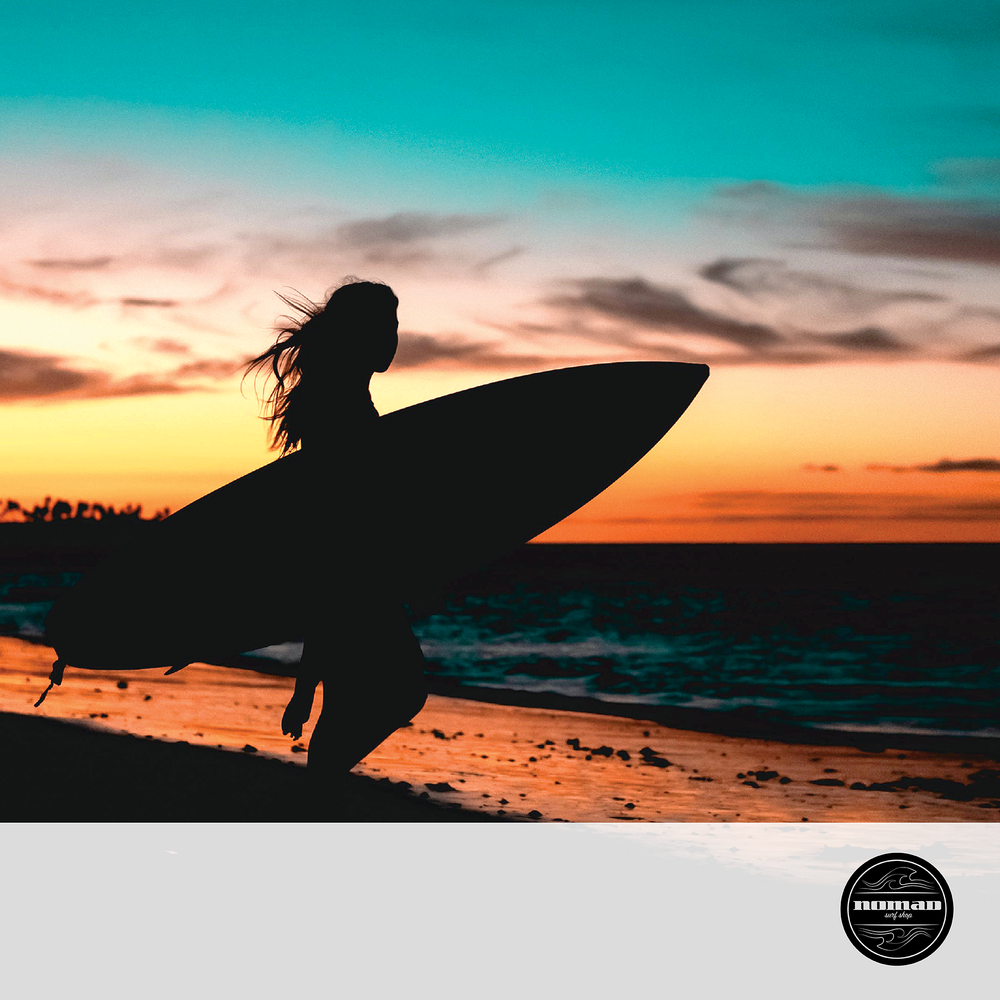A girl holding a surfboard looks out over the ocean as the sun sets