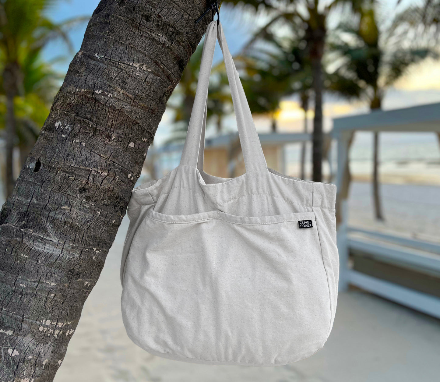 A Sonoma bag hangs peacefully from a palm tree. Behind it lies a tropical beach at sunrise, dotted by Bali beds.