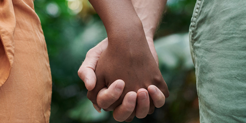 A light-skinned man holds the hand of a woman of color. In the background, is the blurry pattern of tropical leaves. The photo is cropped to show only that much information.