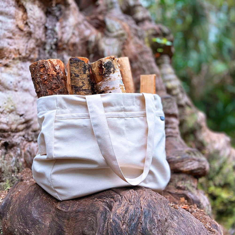 An Olive+Comet Sonoma bag, stuffed with firewood, sits upon the overgrown root of an enormous tree. The blurred background reveals woodland & green patterns.