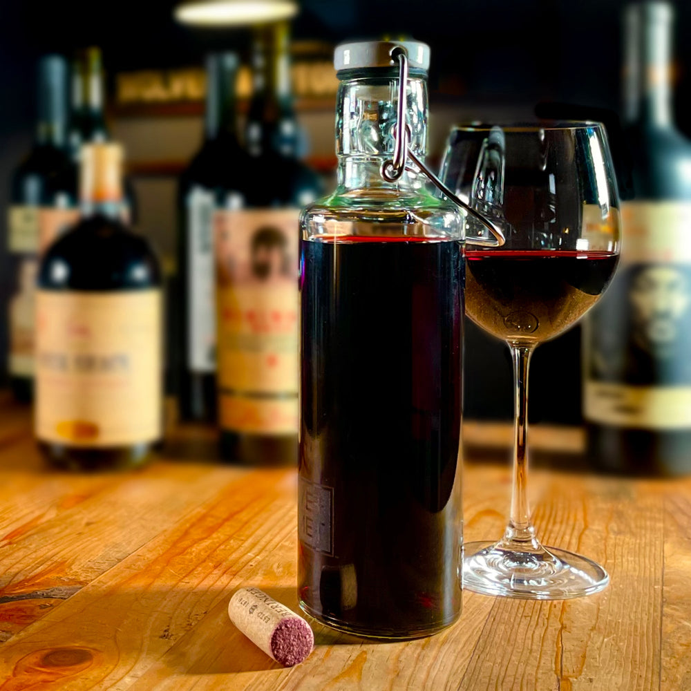 The Olive+Comet Hadley bottle sits, filled with red wine, next to a cork and a partially filled wine glass. The background is blurry, but composed of various wine bottles.