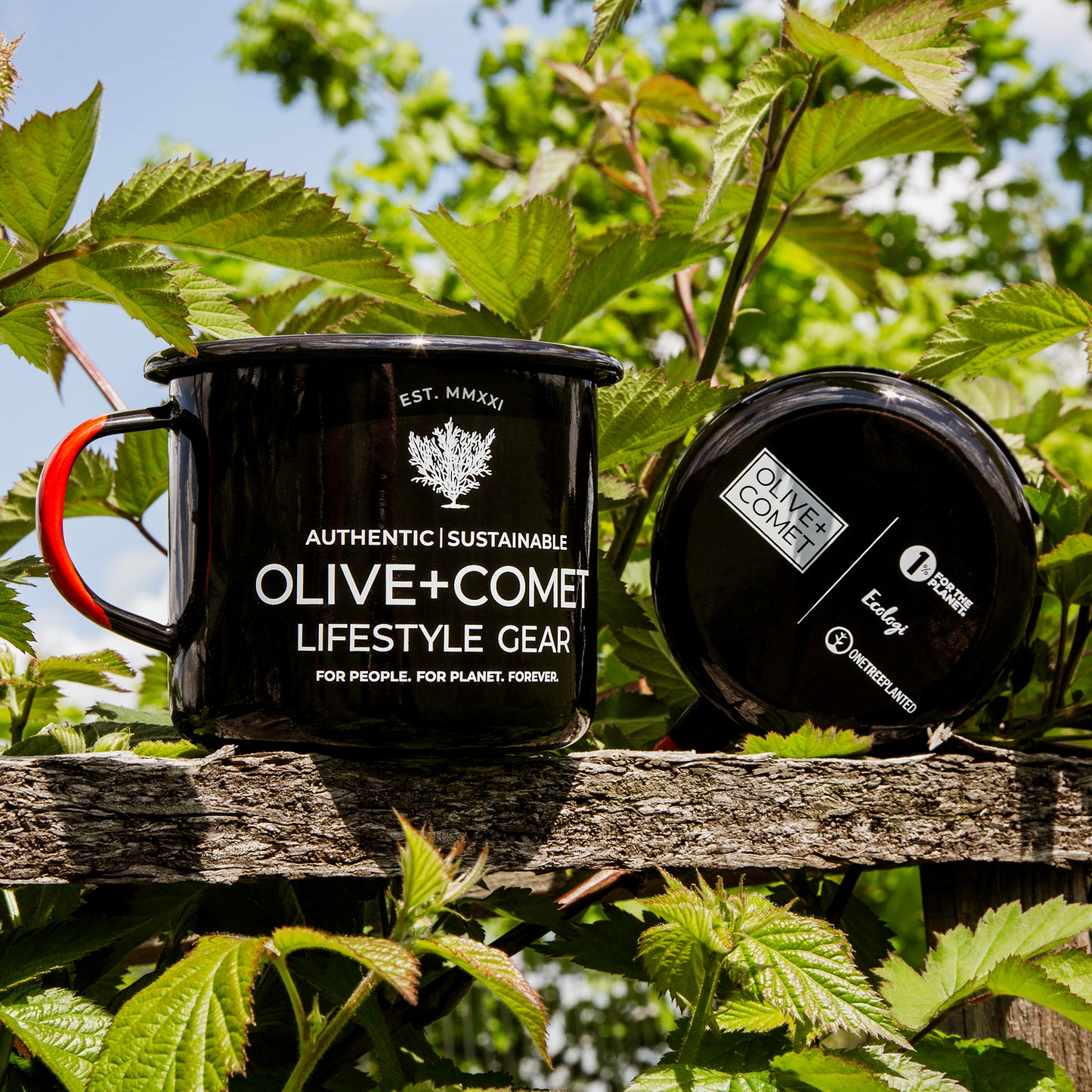 A pair of black Kambi mugs sits on a fence surrounded by green leaves