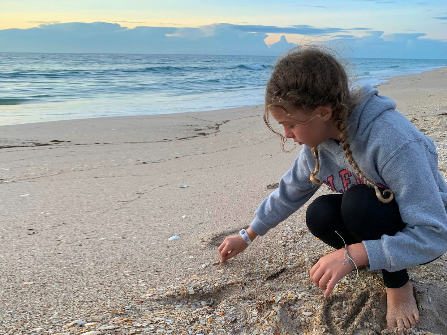 A young girl (the author's youngest daughter) sits at the beach—engrossed in the "art" she is creating in the sand as she draws with a shell. In the background, the sun sets over the ocean.